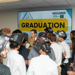 Students in welding helmets stand before a sign that reads "graduation."