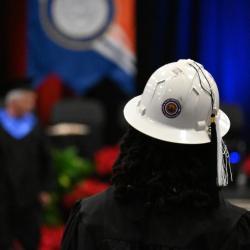 A woman with black curly hair walks down a graduation aisle wearing a hard hat.