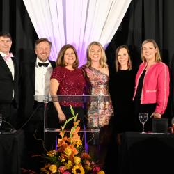 Eight people in formal wear stand in spotlights on a stage during Dinner of Firsts 2026.