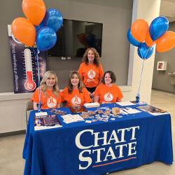 Four women in orange shirts sit behind a table with a ChattState tablecloth and blue and orange balloons.