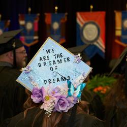 A student attending graduation wears a mortarboard that reads "We Are the Dreamers of Dreams."
