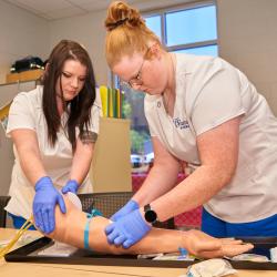 Two female nursing students in white scrubs practicing inserting an IV needle into a plastic prosthetic arm.