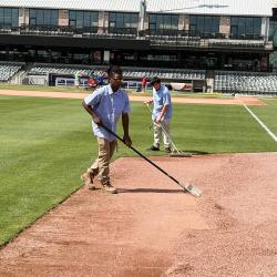 Two male students in workware use rakes to even out gravel on a baseball field.