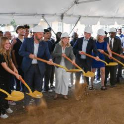 A line of people in hard hats toss dirt with ceremonial shovels. A line of people in hard hats toss dirt with ceremonial shovels.