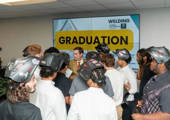 Students in welding helmets stand before a sign that reads "graduation."