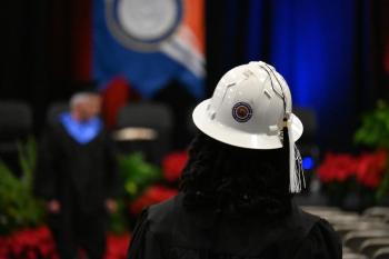 A woman with black curly hair walks down a graduation aisle wearing a hard hat.