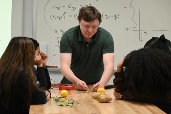 Students look on as an instructor attaches an LED light to a potato and lemon to demonstrate nuclear power. Students look on as an instructor attaches an LED light to a potato and lemon to demonstrate nuclear power.