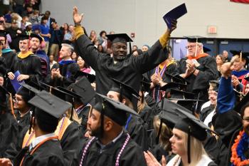 A Chattanooga State graduation in regalia stands with his arms raised surrounded by fellow graduates. A Chattanooga State graduation in regalia stands with his arms raised surrounded by fellow graduates.