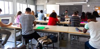 A group of students in a classroom face a teacher and blackboard with electrical diagrams on it.