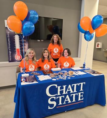 Give Day 2026-2 Four women in orange shirts sit behind a table with a ChattState tablecloth and blue and orange balloons.