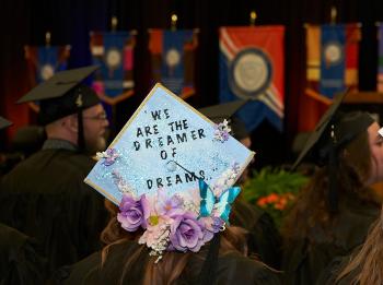 A student attending graduation wears a mortarboard that reads "We Are the Dreamers of Dreams." A student attending graduation wears a mortarboard that reads "We Are the Dreamers of Dreams."