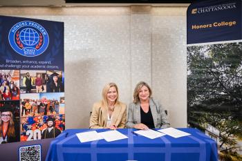 Honors Articulation Two blonde women in business casual clothing sign articulation agreements on a blue table flanked by signs promoting ChattState and UTC.