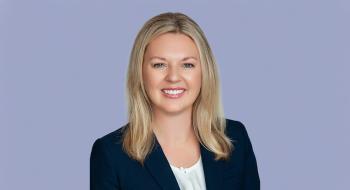 Tiffanie Robinson, a blonde woman in a black coat, smiles in front of a light blue background.