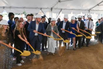 A line of people in hard hats toss dirt with ceremonial shovels. A line of people in hard hats toss dirt with ceremonial shovels.