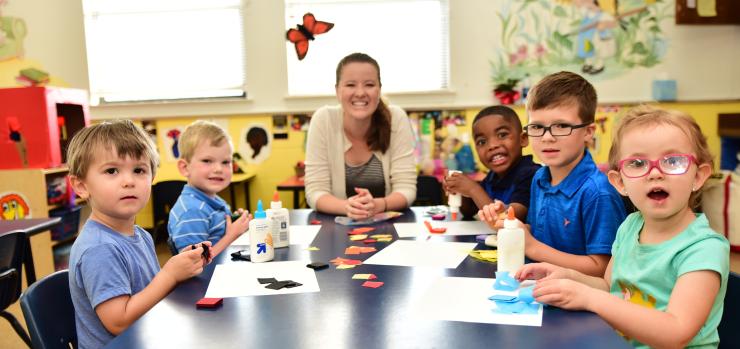 pre-k classroom scene with smiling female teacher and five children sitting together at a table