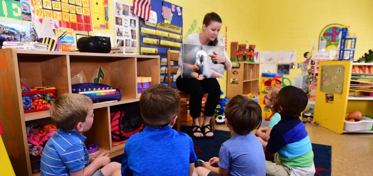 pre-k classroom scene with female teacher reading to five children sitting in the floor
