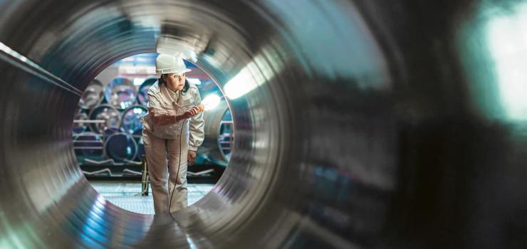 a woman wearing a hard hat shining a light inside of a large pipe to inspect it